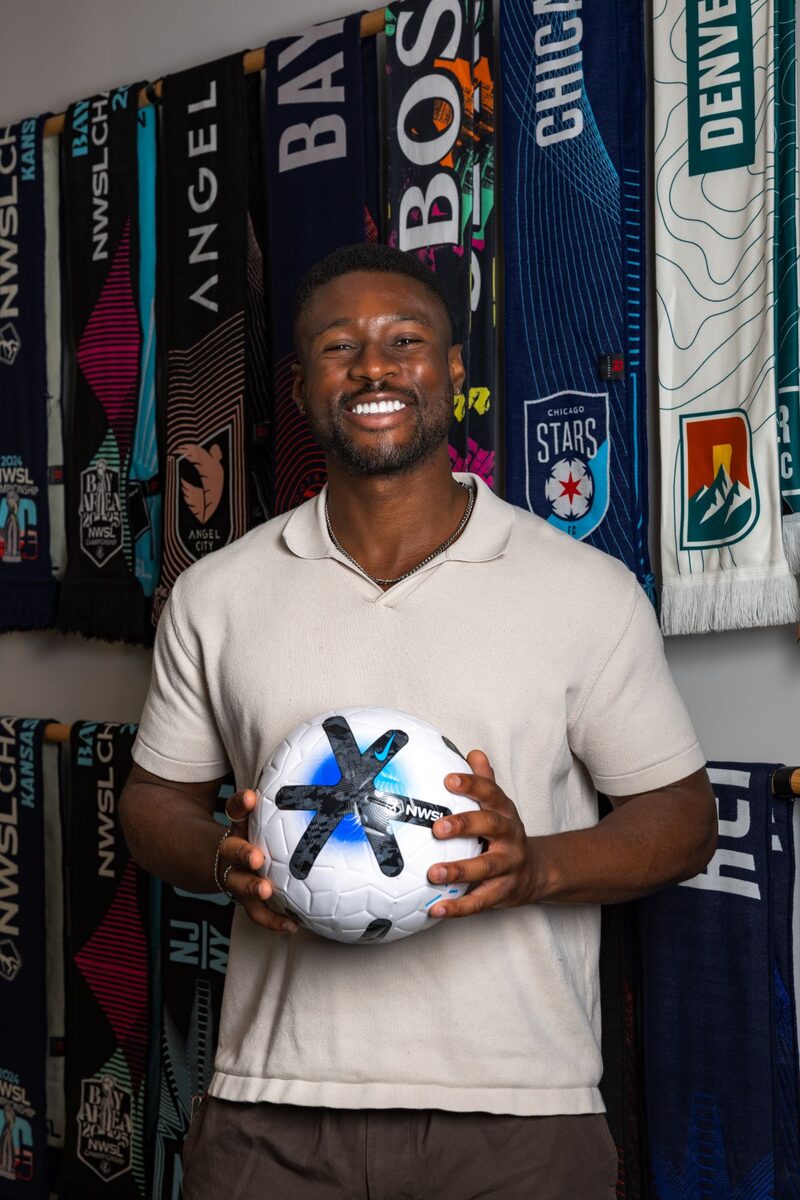 LJ Umunna smiling and holding a soccer ball in front of a wall of NWSL club scarves